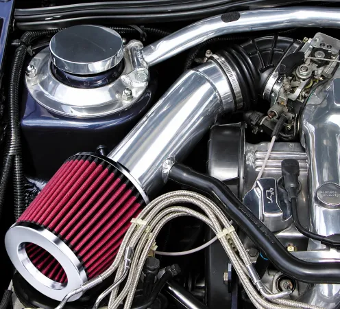 Een rood, open sportluchtfilter in de motorruimte, verbonden met een glanzend zilveren cold air intake-buis naar het motorblok. Omringd door zilveren motoronderdelen, zwarte slangen en gevlochten leidingen valt het filter extra op.