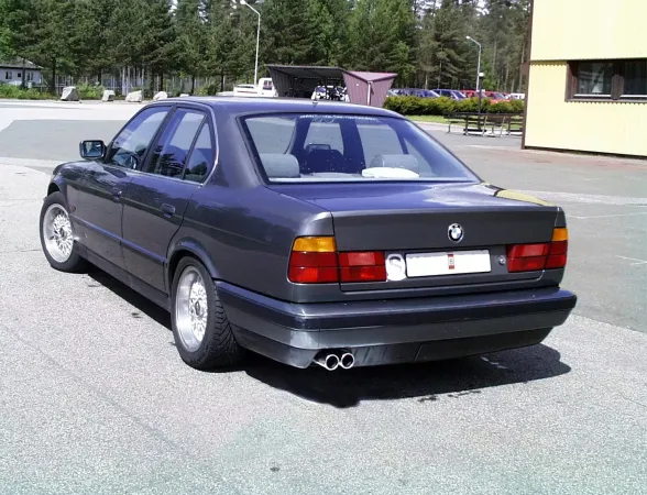 Achteraanzicht van een donkergrijze BMW E34. Je ziet het chroomstalen duplex sportuitlaatsysteem met 2x70 mm uitlaatpijpen, gemonteerd onder de bumper. De auto staat geparkeerd op een parkeerplaats, met bomen en een gebouw op de achtergrond.