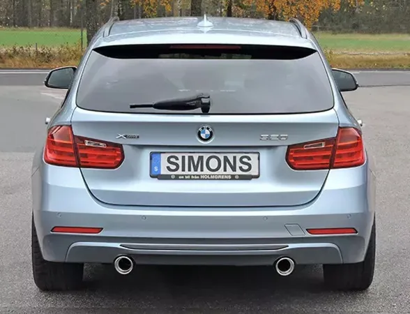 Achteraanzicht van een lichtblauwe BMW F31 320i estate met duplex sportuitlaatsysteem. Nummerplaat met Simons belettering. De uitlaatpijpen van het uitlaatsysteem zijn rechts en links geplaatst. Achterlichten en BMW logo zijn zichtbaar.